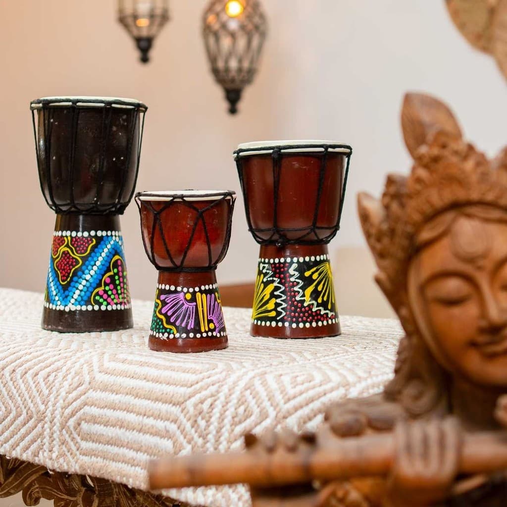 Três tambores djembê de madeira importados de Bali, em cores marrom, preto e vermelho, dispostos em uma mesa com fundo de parede clara e estátua de Buda.