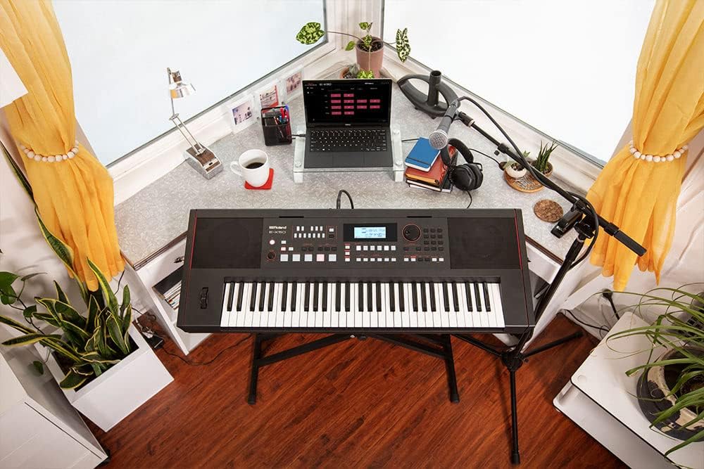Teclado Roland E-X50 em suporte com microfone, laptop e plantas em sala iluminada