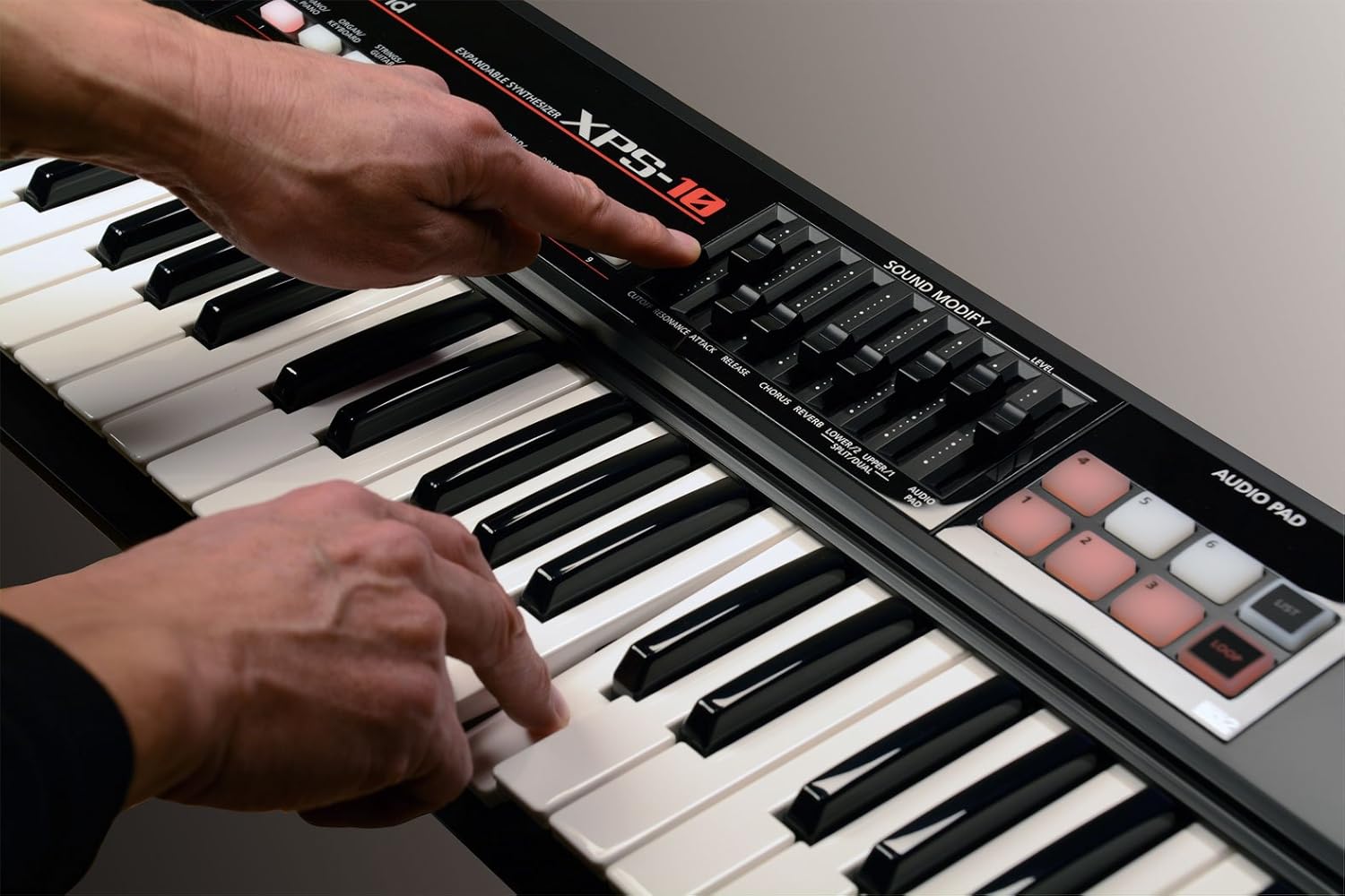 Mãos tocando teclado Roland XPS-10 preto com controle sliders e pads áudio.
