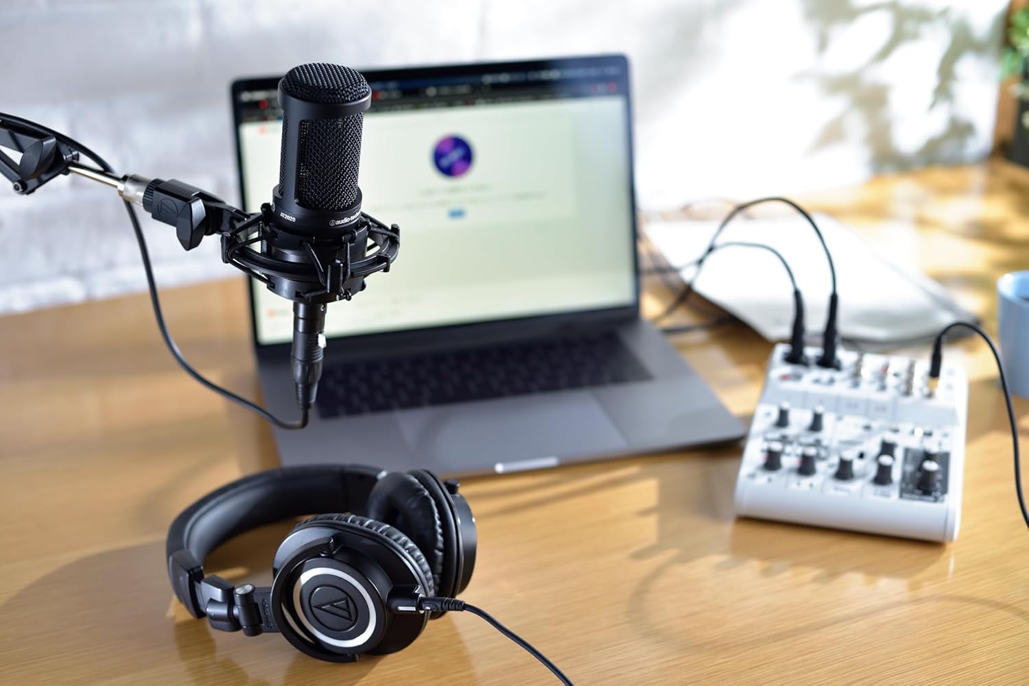 Microfone condensador cardioide Audio-Technica AT2020 em mesa de gravação com notebook, mixer e fones de ouvido
