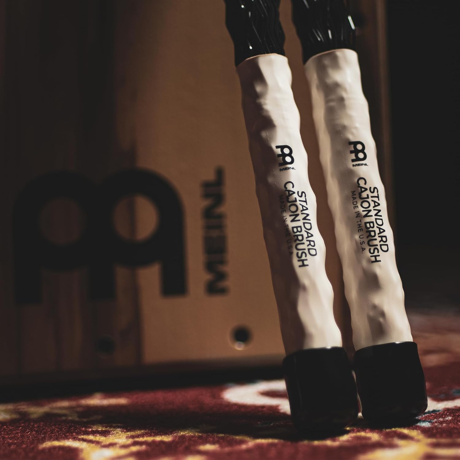 Escovas Meinl SB305 para cajon em pé sobre tapete vermelho com caixa da marca ao fundo.