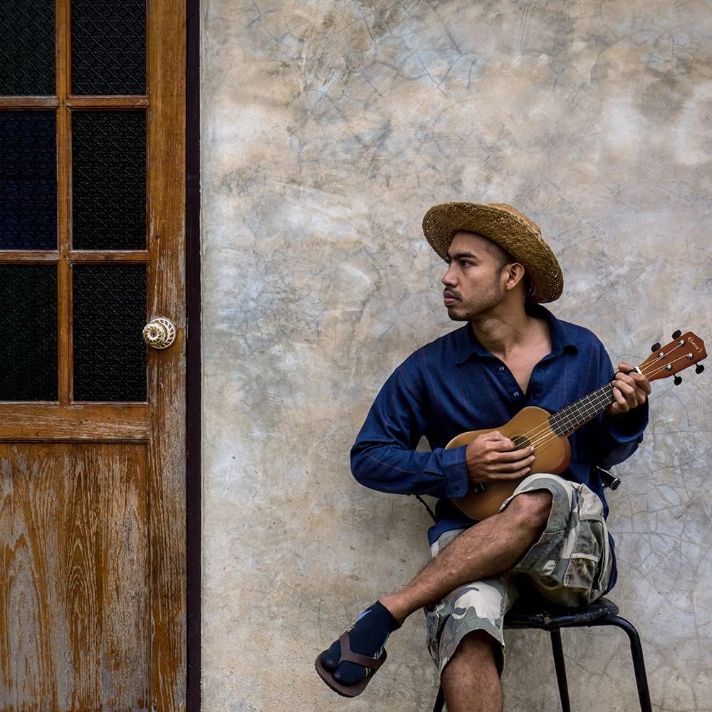 Homem tocando ukulele acústico soprano Queen's 21 polegadas sentado em banco, com parede e porta ao fundo.