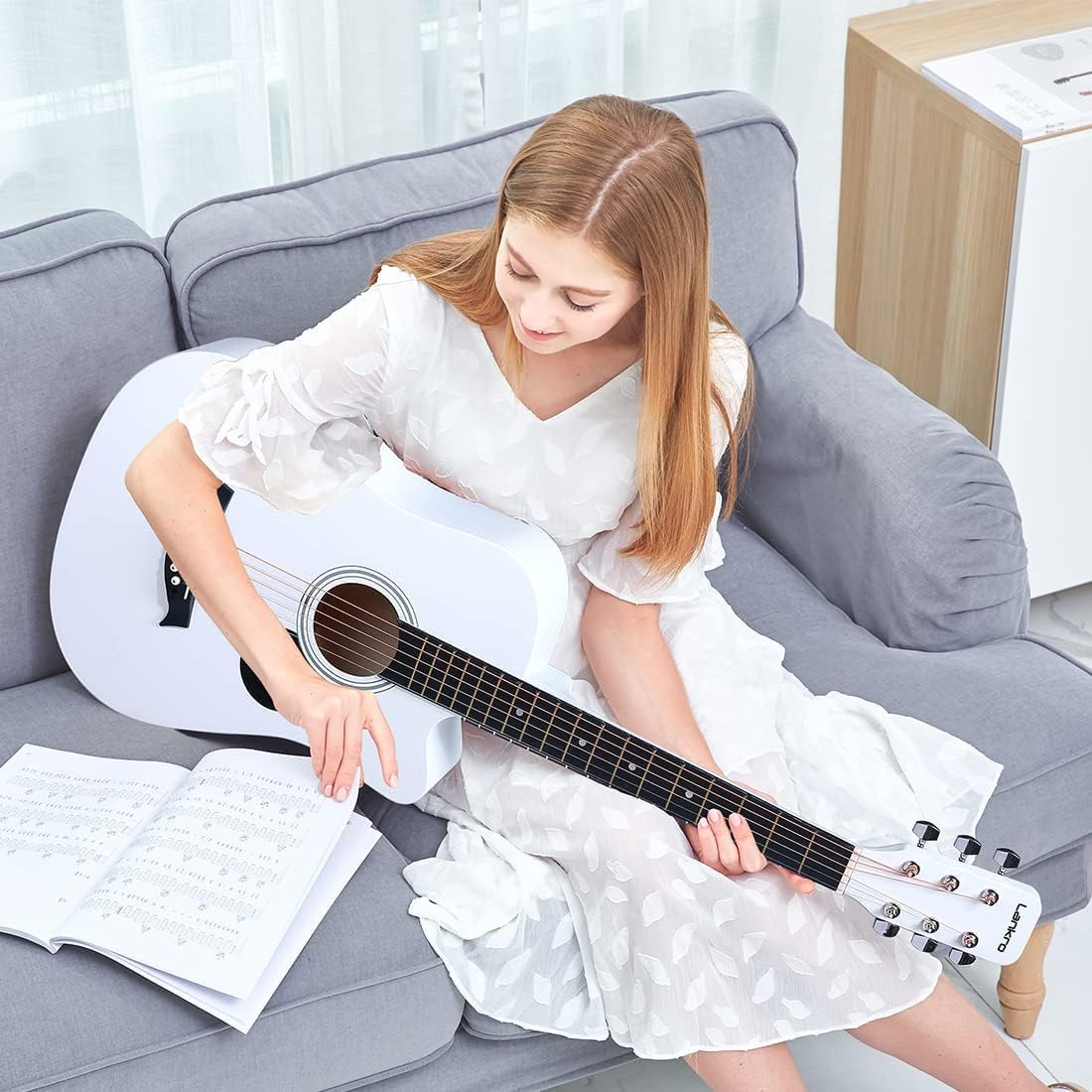 Mulher tocando guitarra acústica branca Lankro sentada em sofá cinza com livro de partituras