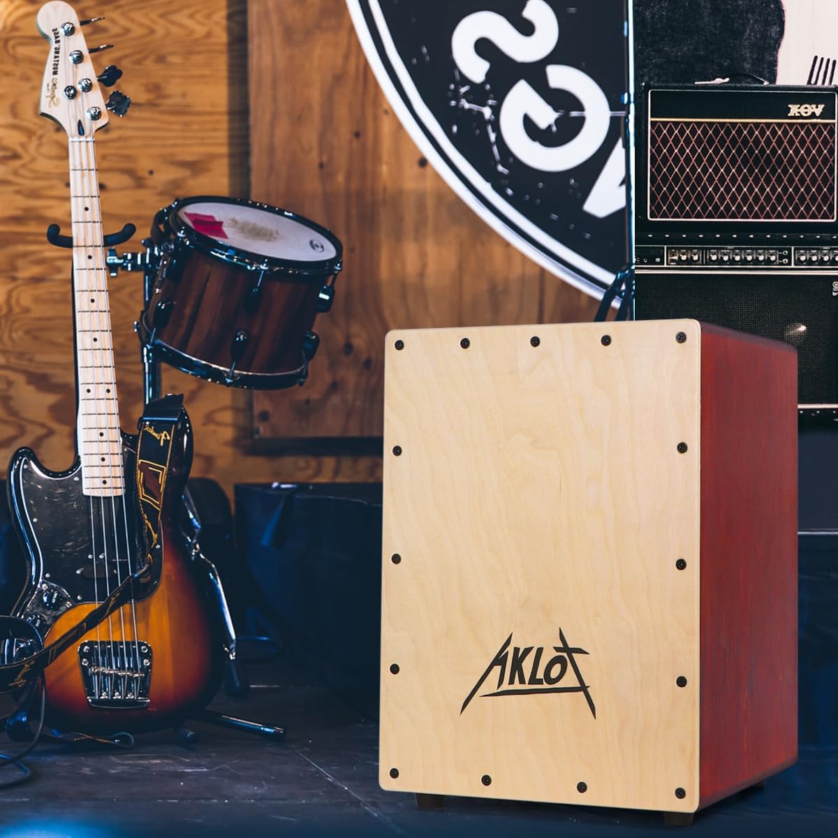 Cajon AKLOT em madeira de bétula em ambiente de palco com baixo elétrico e tambor ao fundo.
