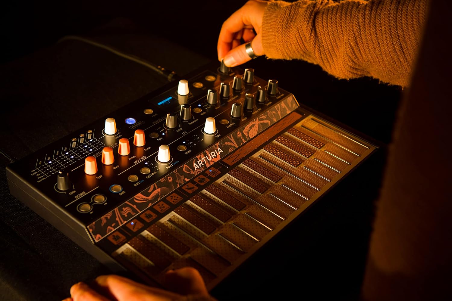 Arturia MicroFreak sintetizador híbrido em ambiente escuro com iluminação amarela, mão ajustando knob e teclado PCB sensível à pressão.