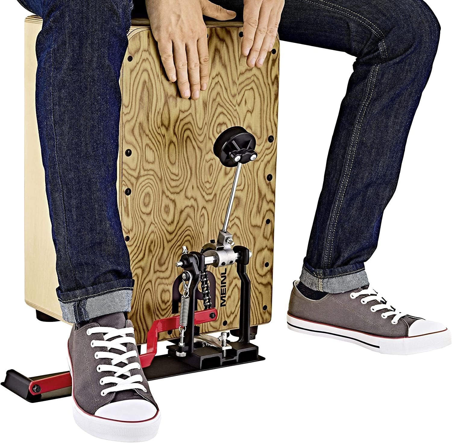 Pessoa tocando cajon com pedal Meinl ativado pelo calcanhar, estrutura preta e vermelha, com tênis cinza.