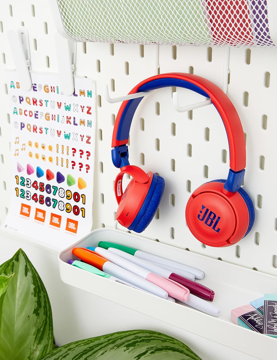 Fone de ouvido infantil JBL JR310BT vermelho com azul pendurado em suporte branco próximo a material escolar.