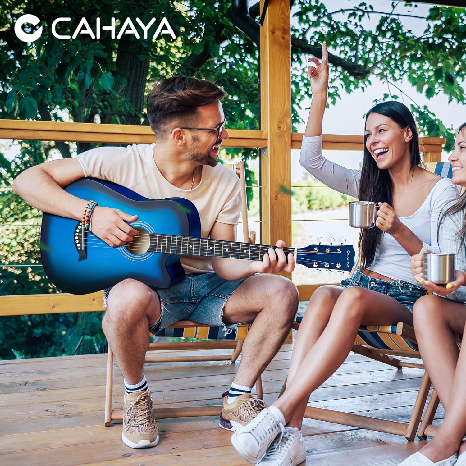 Homem tocando guitarra acústica CAHAYA azul fosco ao ar livre com duas mulheres sorrindo ao lado.