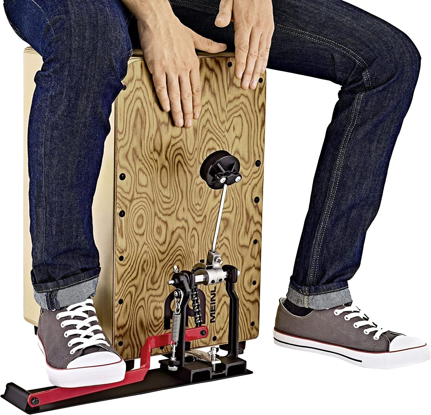 Pessoa usando pedal Meinl para cajon ativado pelo calcanhar, pedal preto e vermelho, cajon madeira clara, jeans e tênis cinza.
