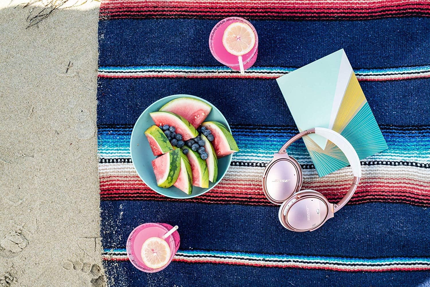 Fones de ouvido Bose QuietComfort 35 II rosa sobre toalha de praia com frutas, bebidas e livro.