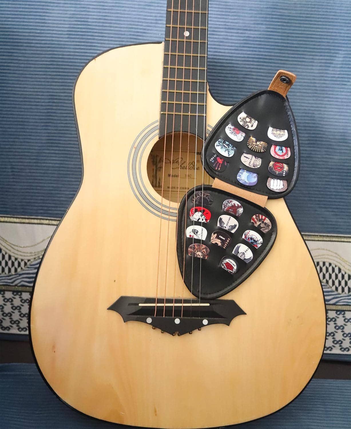 Estojo triangular para palhetas em couro sintético marrom aberto com palhetas coloridas ao lado de guitarra acústica clara.