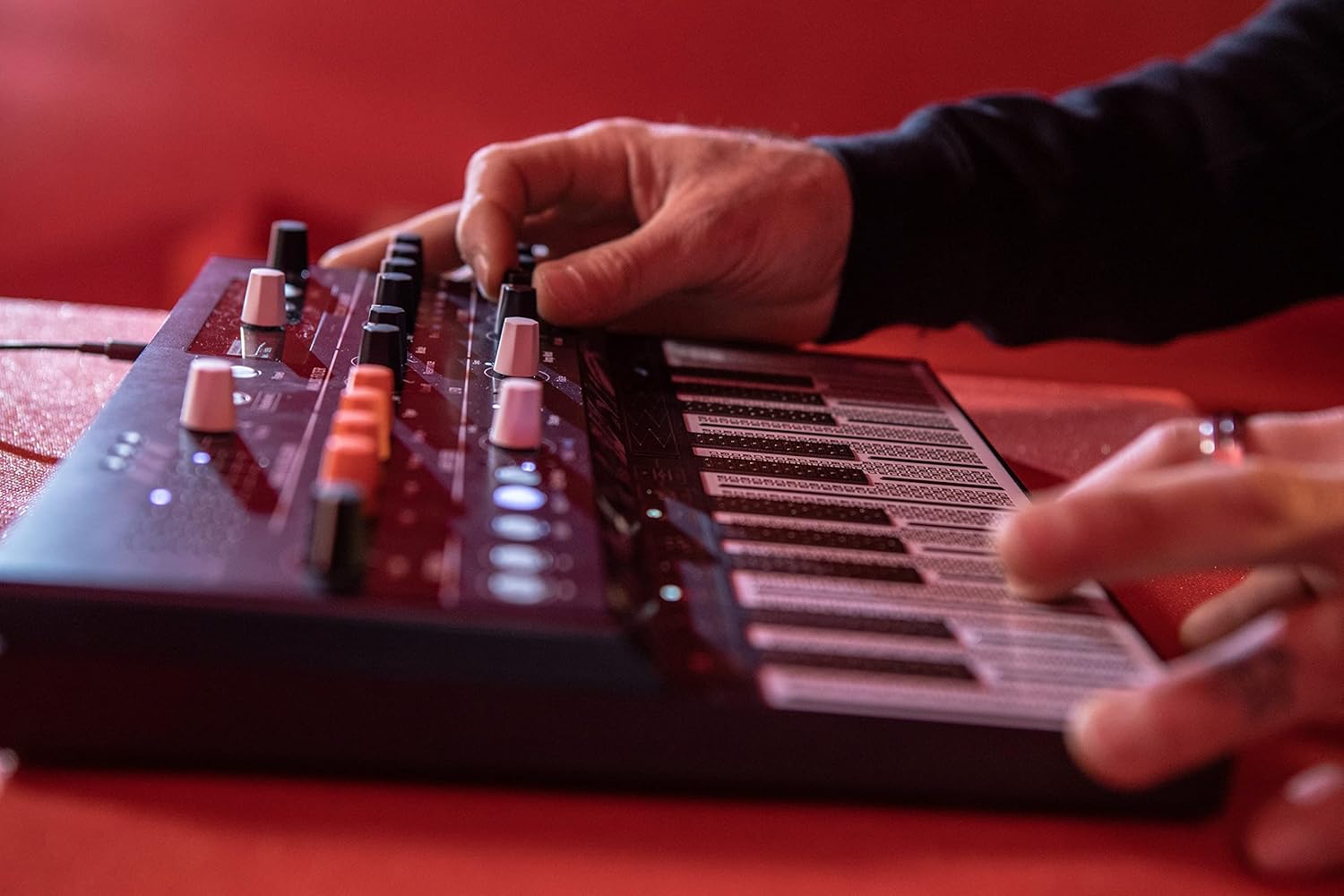 Lado do sintetizador Arturia MicroFreak com mãos ajustando knobs e teclado PCB sensível à pressão iluminado em vermelho.