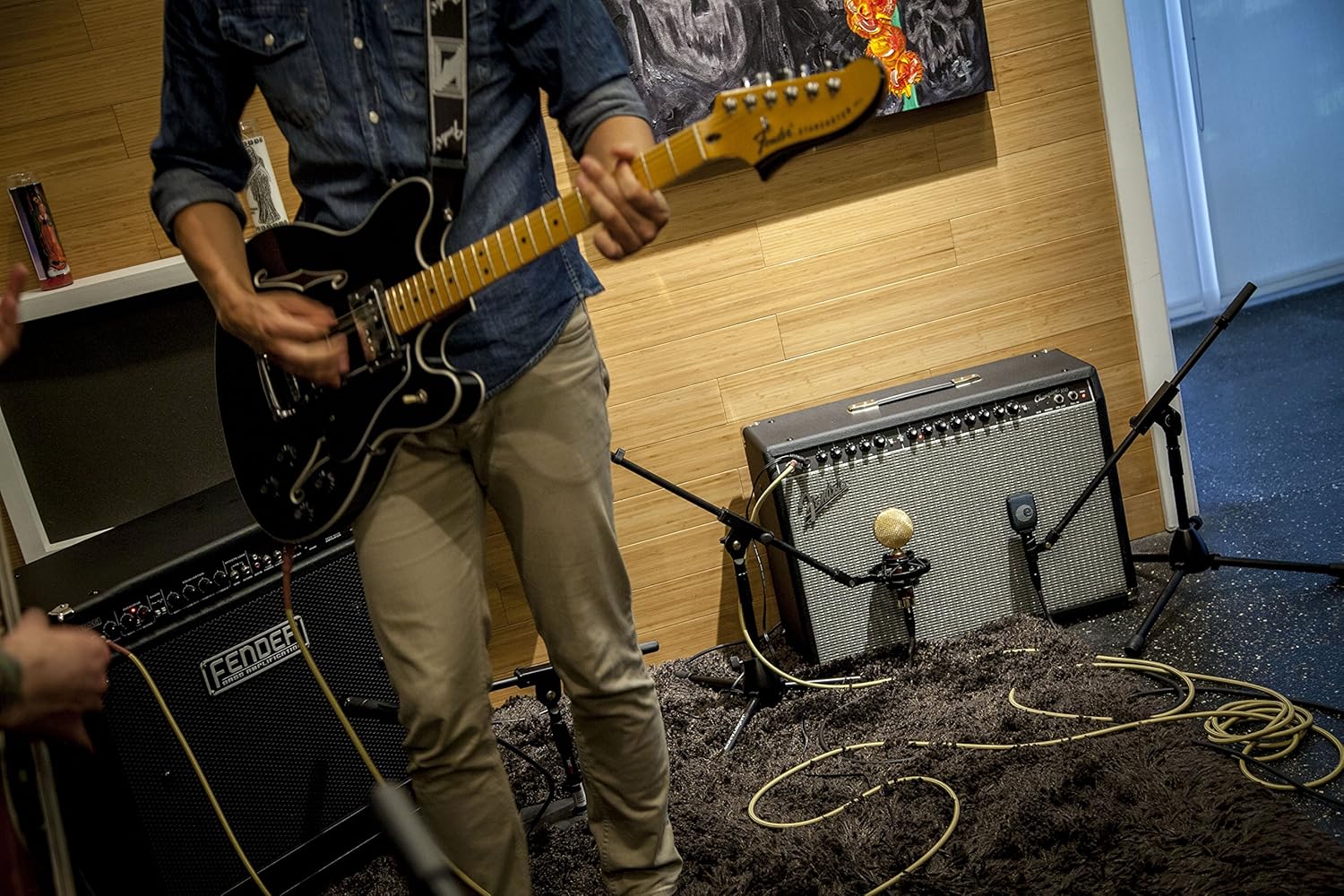 Músico tocando guitarra elétrica Fender em estúdio com amplificador combo Fender Champion 100 conectado e microfones para captação.