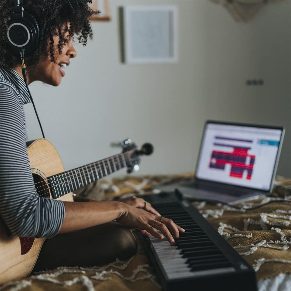 Mulher tocando teclado com guitarra acústica no colo em ambiente doméstico com laptop e fones de ouvido.