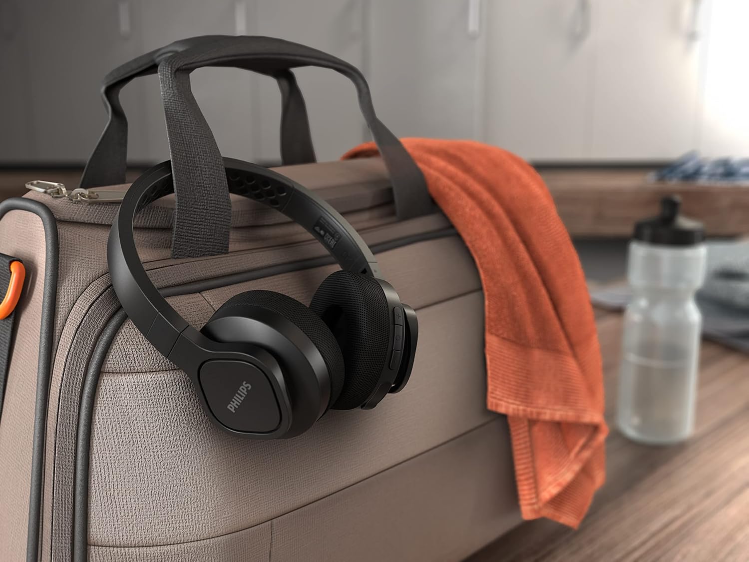 Fone de ouvido Philips preto pendurado em mala cinza com toalha laranja em fundo desfocado